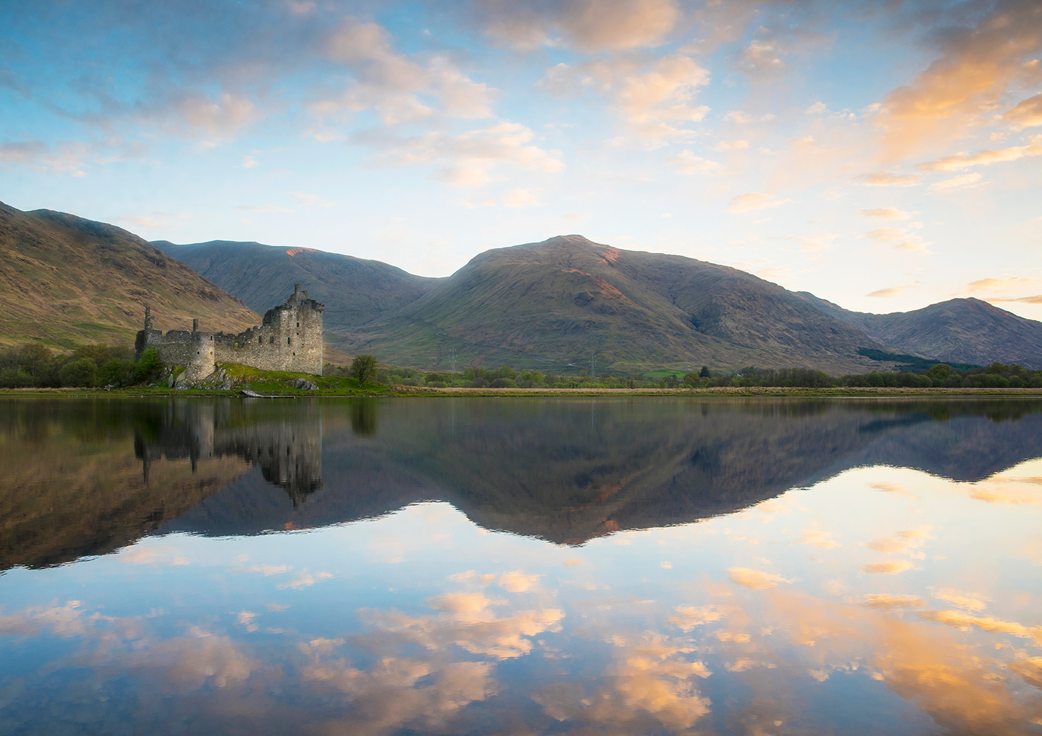 A caslte ruin is reflected on a calm loch, with hills beyond